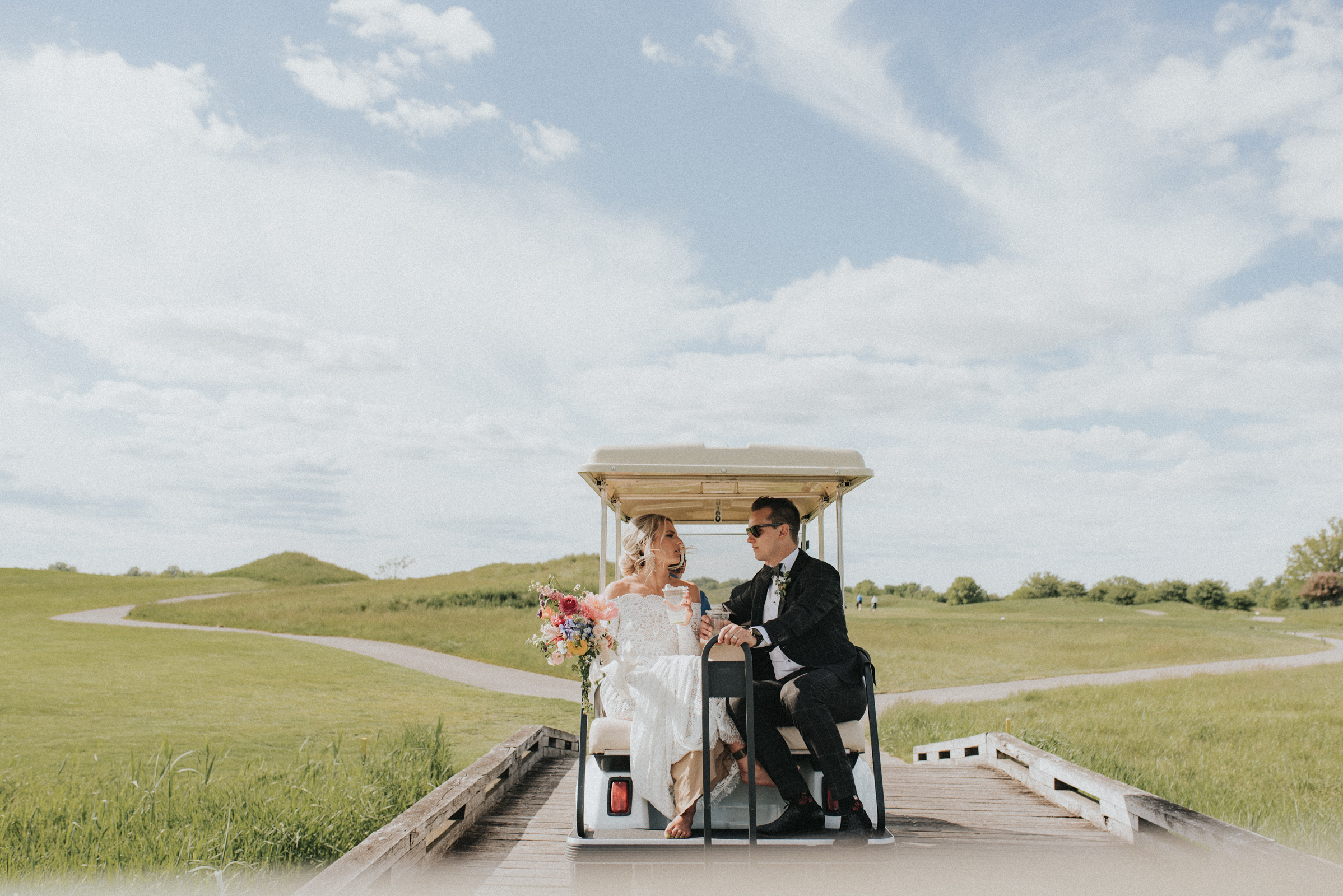 Couple riding in a Golf cart on their wedding day with the greens of Piper's Heath Golf Club in the background.