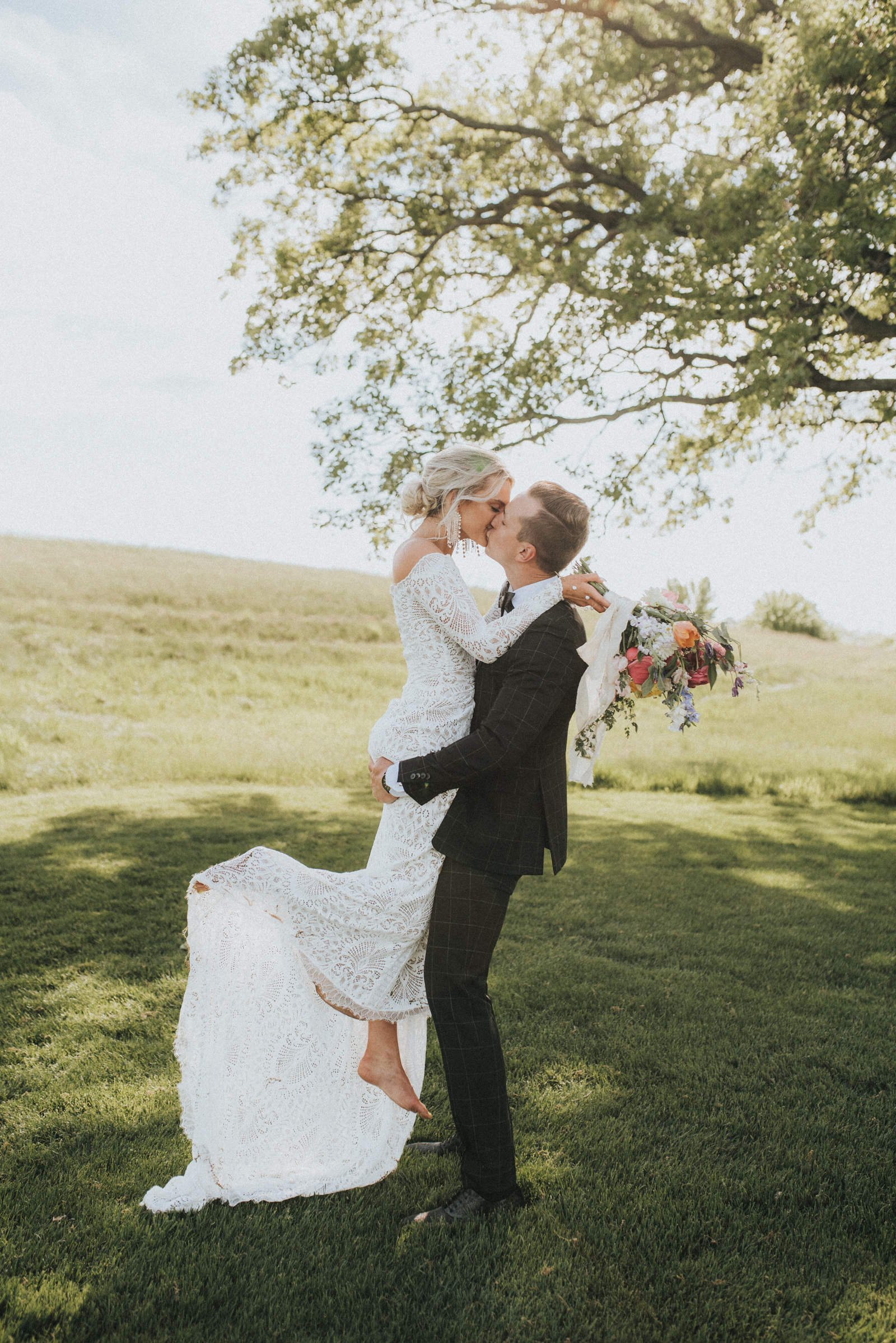 Groom holding barefoot Bride while kissing her with rolling hills in the background.